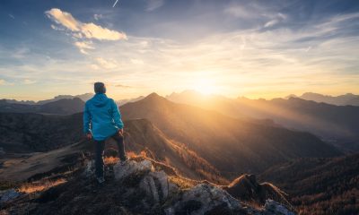 Man on stone on the hill and beautiful mountain valley in haze at colorful sunset in autumn. Dolomites, Italy. Guy, mountain ridges in fog, orange grass and trees, blue sky with sun in fall. Hiking