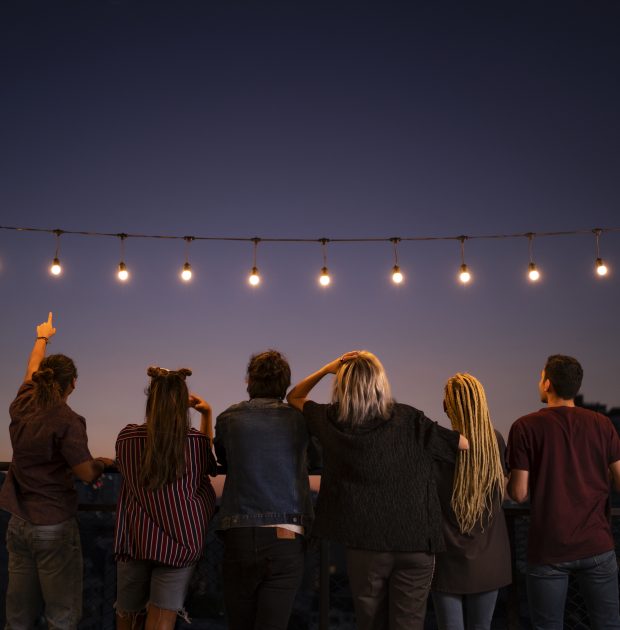 Friends spending leisure time under string lights on rooftop at dusk